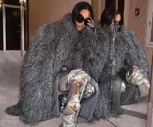 Person wearing a silver textured coat and metallic thigh-high boots crouching inside a hallway with dark sunglasses.