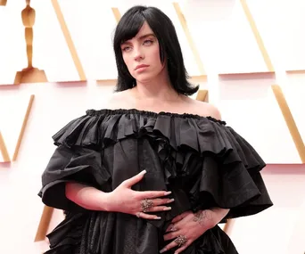 Billie Eilish at the Oscars in a black off-the-shoulder ruffled dress, standing on a red carpet.