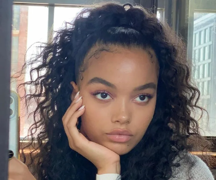 Woman with curly hair and makeup, resting her chin on her hand, looking pensive in natural light.