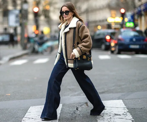 Woman in a shearling jacket and jeans crossing a city street, carrying a black handbag and wearing sunglasses.