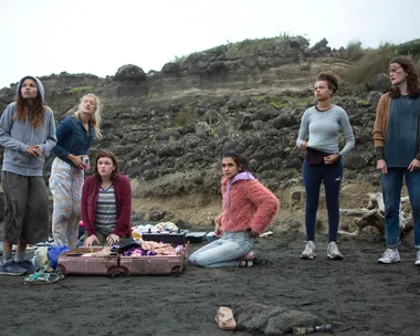 A group of girls on a rocky beach, surrounded by luggage and clothes, looking concerned. Scene from "The Wilds."