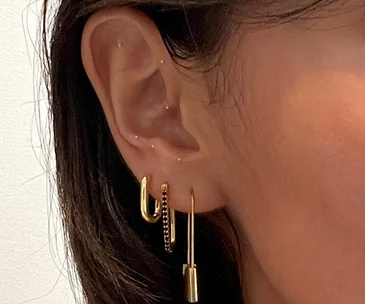 Close-up of an ear with gold ear seeds and multiple earrings, including hoops and dangle earrings.