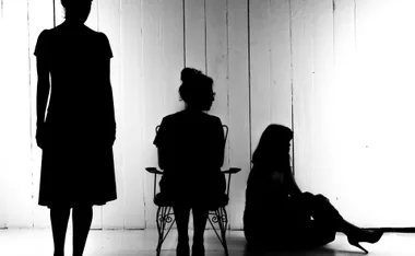 Silhouette of three women, one standing, one seated, and one sitting on the floor, against a wooden wall backdrop.