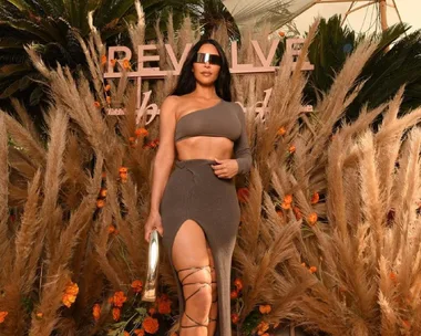 Person in brown asymmetric outfit and sunglasses at Revolve Festival, surrounded by pampas grass and flowers.