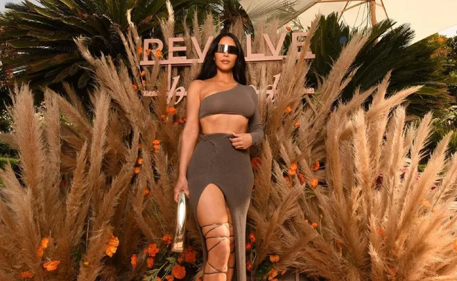 Person in brown asymmetric outfit and sunglasses at Revolve Festival, surrounded by pampas grass and flowers.