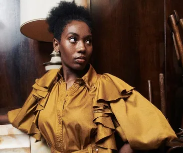 A person in a mustard ruffle dress stands against a wooden wall, under a lamp, looking to the side with a serious expression.