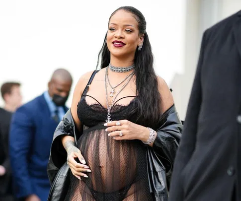 Pregnant woman in stylish black outfit and jewelry, smiling at an event with people in the background.