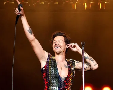 Harry Styles performing on stage, smiling and holding a microphone, wearing a sequin outfit.