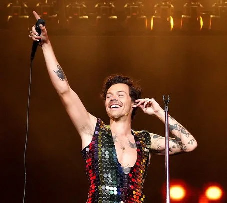 Harry Styles performing on stage, smiling and holding a microphone, wearing a sequin outfit.