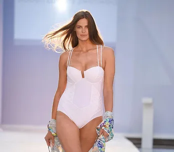 Model in a white swimsuit walks the runway at Australian Fashion Week, holding a colorful scarf.