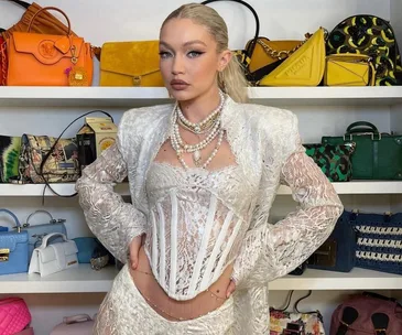 A woman in a lace outfit and pearl necklace poses in front of a shelf filled with colorful handbags.