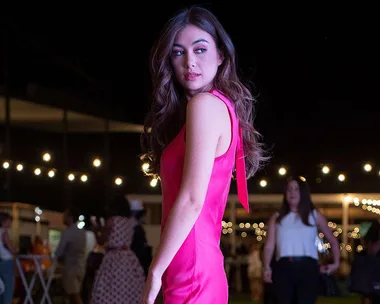 A woman in a pink dress stands at a nighttime event with string lights in the background.