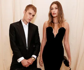 A couple poses in formal evening wear, the man in a tuxedo and the woman in a black strapless gown, standing against a beige background.