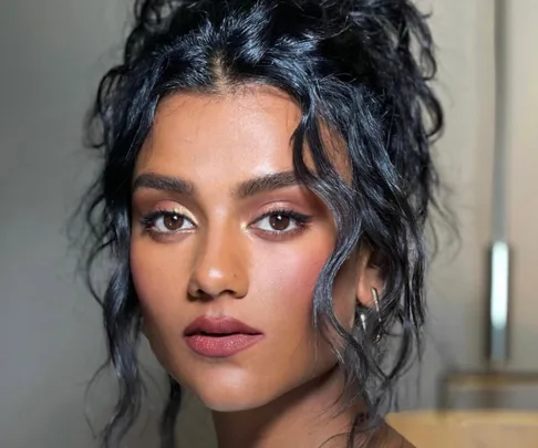 A person with curly dark hair and natural makeup, wearing hoop earrings, looking directly at the camera.