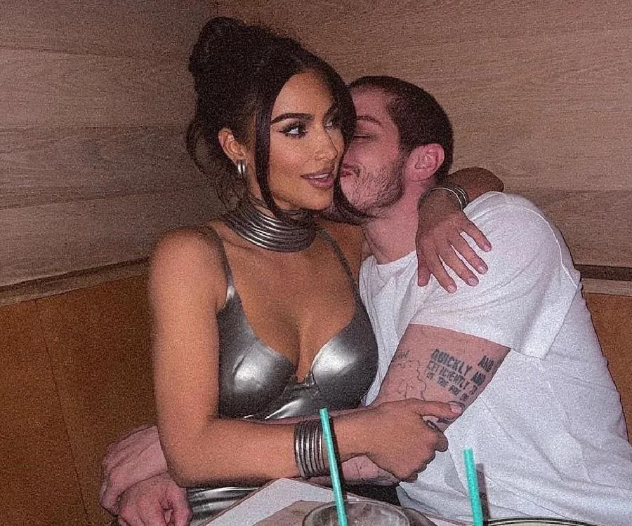 A woman in a metallic dress and a man in a white t-shirt are embracing in a cozy environment.