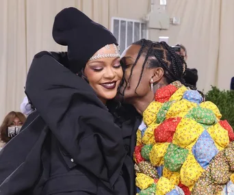 Two people embrace warmly; one wears a dark outfit with a headpiece, the other in a colorful jacket, sharing a tender moment.