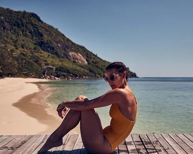 Woman in orange swimsuit sitting on a wooden deck by a tropical beach and clear blue ocean.