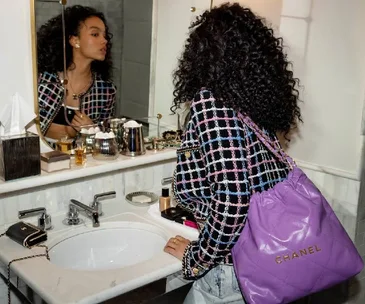 Woman in checkered jacket with a purple Chanel bag looks in a bathroom mirror, surrounded by luxury cosmetics.