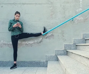 Woman stretching on stairs while using a smartphone, wearing headphones and a green jacket.