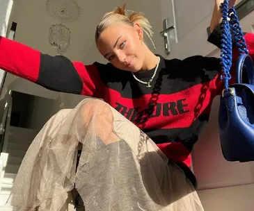 Woman in red and black "The Cure" sweater with tulle skirt, holding a blue purse, sits on stairs in sunlight.