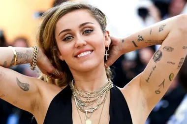 Woman smiling with arms raised, showcasing numerous tattoos on her arms and wearing layered necklaces and earrings.