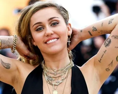 Woman smiling with arms raised, showcasing numerous tattoos on her arms and wearing layered necklaces and earrings.