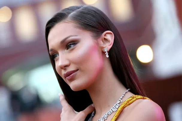 A person with long dark hair and diamond jewelry looks away, set against a blurred background.