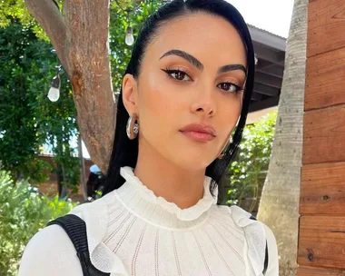 A woman with long black hair and strong eyebrows, wearing a white top, stands outdoors near trees and a wooden wall.