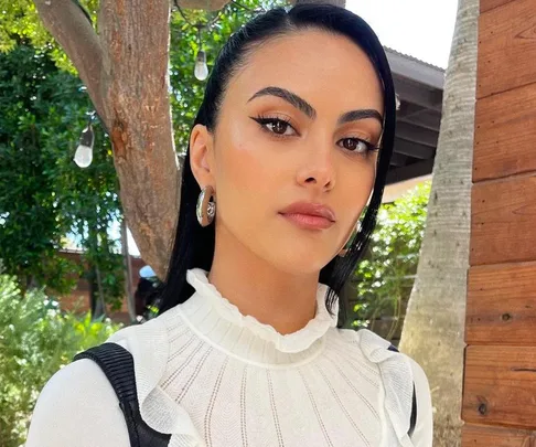 A woman with long black hair and strong eyebrows, wearing a white top, stands outdoors near trees and a wooden wall.