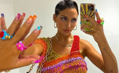 A person takes a mirror selfie, showcasing colorful nails, rings, and a vibrant crocheted top with a phone case design.