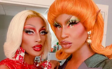 Two drag queens in vivid makeup and wigs, one with blonde hair, the other with orange, pose together backstage.