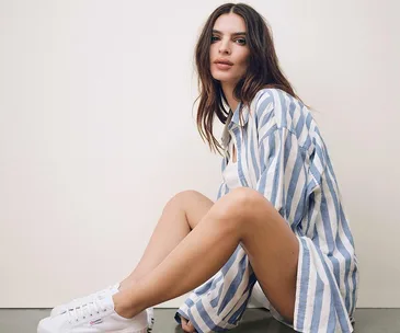 Woman in a blue striped shirt and white sneakers sitting on the floor, posing against a simple backdrop.