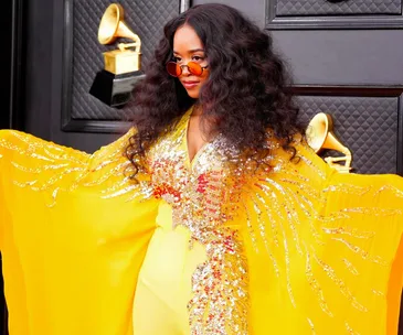 A person in a sparkly yellow outfit and round sunglasses poses on the Grammys 2022 red carpet with Grammy trophies in the background.