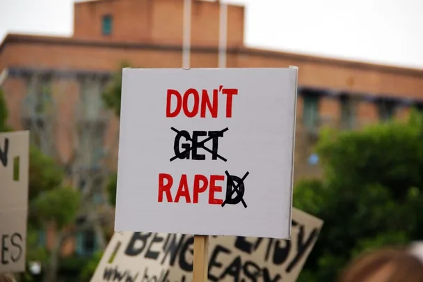 Protest sign reading "DON'T GET RAPED" with "GET" crossed out, held outdoors.