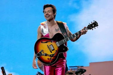 Singer performing on stage, smiling, wearing pink outfit, and playing a guitar against a blue sky background.