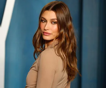 Woman with long wavy hair in a beige outfit, posing against a blue background.