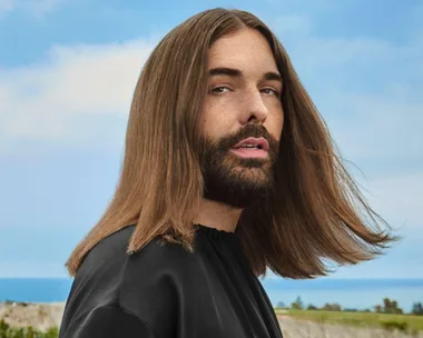 Person with long hair and a beard, wearing a black top, standing outdoors with a blue sky in the background.