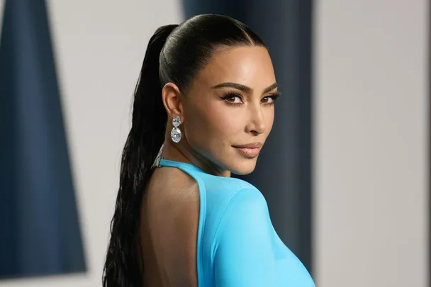 A woman in a sleek blue dress with diamond earrings and a high ponytail, looking over her shoulder at an event.