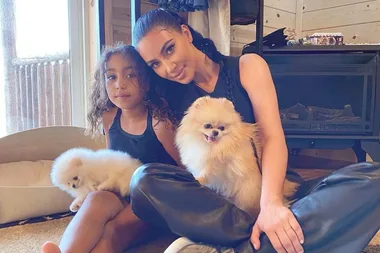 A woman and a young girl sit indoors with two fluffy dogs, smiling and relaxed.