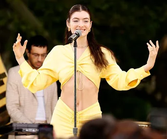 Singer in a yellow outfit performs on stage, smiling with arms raised near a microphone.