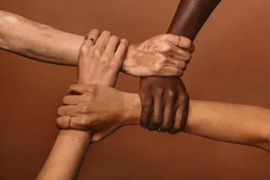 Four hands of diverse skin tones clasp each other's wrists against a brown background, symbolizing unity and support.