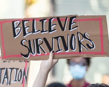 A hand holding a cardboard sign reading "Believe Survivors" during a protest.