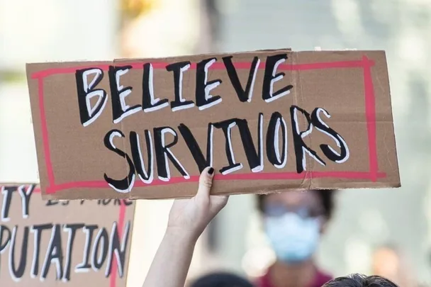 A hand holding a cardboard sign reading "Believe Survivors" during a protest.