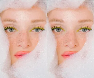 Close-up of a woman's face with freckles, surrounded by soap bubbles, wearing bright eye makeup and pink lipstick.