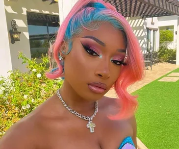 Woman with colorful hair and vibrant makeup poses outdoors, wearing a cross necklace and strapless top.