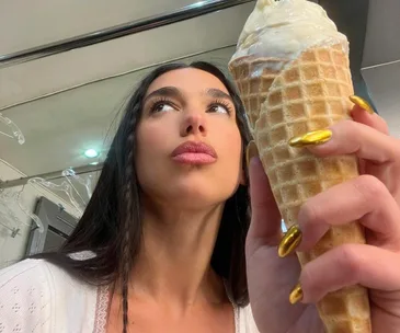 Woman with long hair holds an ice cream cone, showcasing metallic gold nails against a mirrored background.