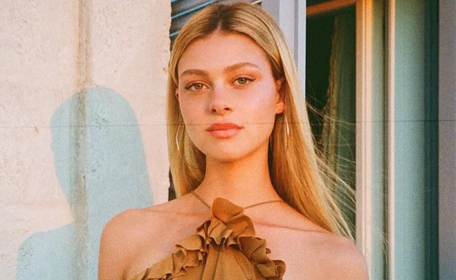 A person with long blonde hair stands in front of a window, wearing a ruffled brown top.