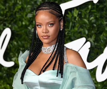 Rihanna in a braided hairstyle, wearing a light teal dress and diamond jewelry, standing against a leafy backdrop.