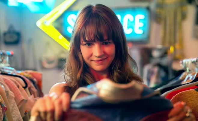 A person smiling while examining clothes in a second-hand shop, with colorful signs in the background.