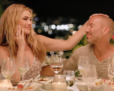 A woman gently touches a man's head at a dinner table, surrounded by dishes and glasses, with city lights in the background.